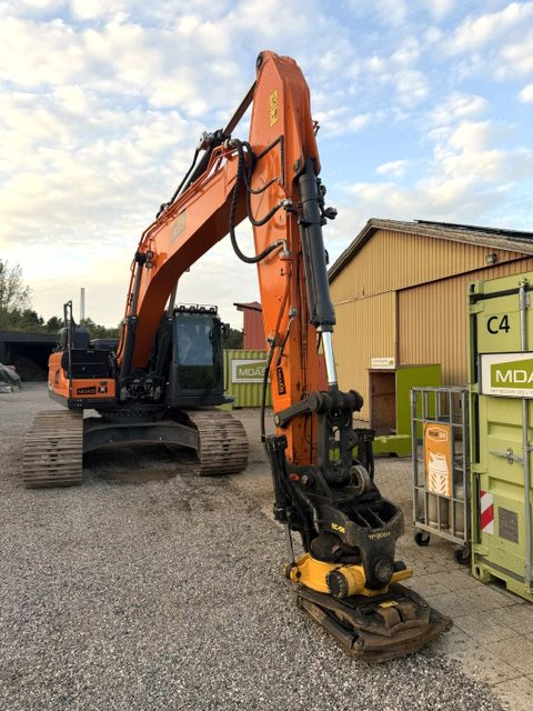Doosan DX 255 LC set forfra i trekvart vinkel med hævet bom og Engcon tiltrotator stillet på gruset foran maskinen, parkeret foran gul hal og grønne containere
