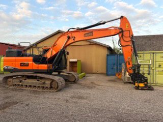 Doosan DX 255 LC set fra venstre side med bommen strakt ud og tiltrotatoren hvilende på jorden, foran gul hal med blå port og grønne containere