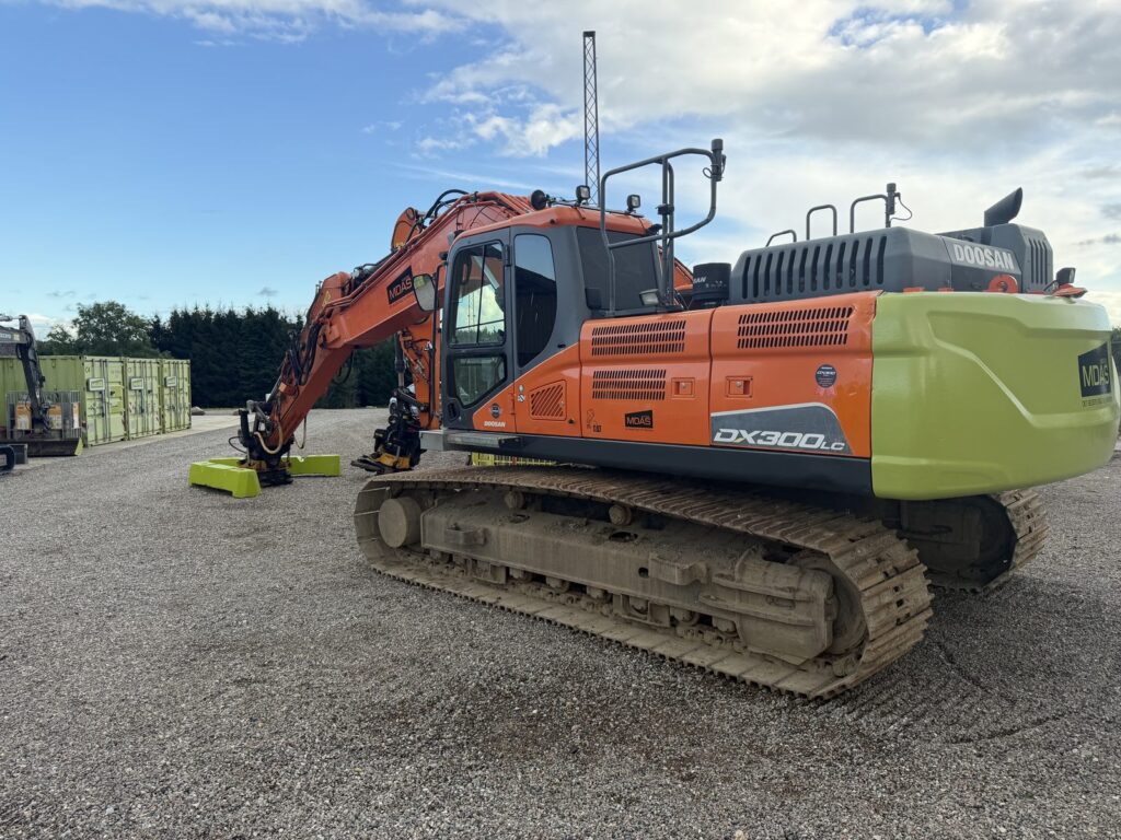 Doosan DX 300 LC bæltegraver set skråt bagfra fra venstre side med bom, Engcon tiltrotator på grønne betonklodser og kabine, parkeret på grusplads