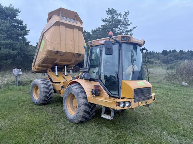 Hydrema 912C dumper set skråt forfra med ladet tippet op, kabinen til højre i billedet, parkeret på græs foran træer