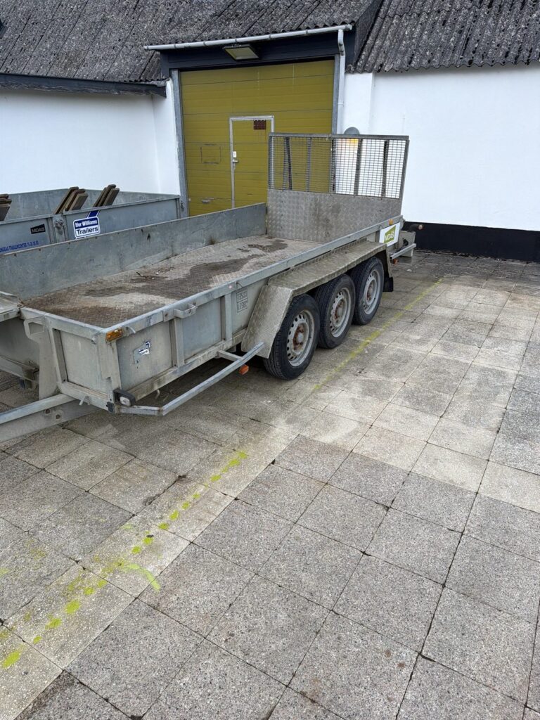 Ifor Williams GP147 maskintrailer set fra siden med tre aksler, alubund og gitterrampe bagtil, parkeret foran en hvid bygning med gul port