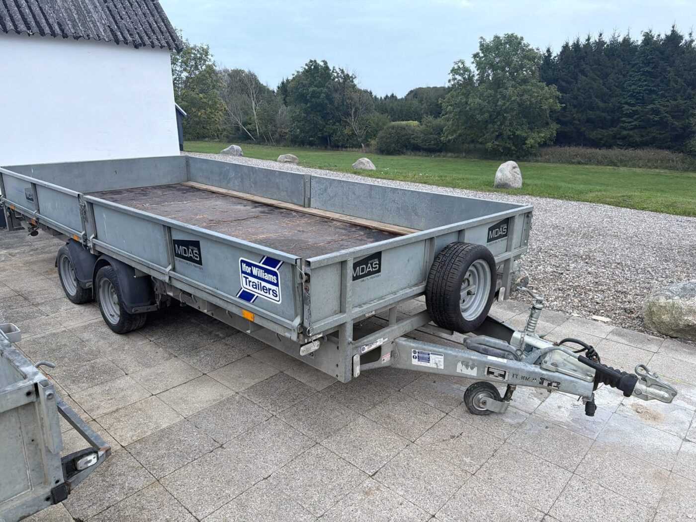 Ifor Williams LM167 trailer set skråt forfra fra højre side med trækstang, reservehjul foran og to aksler, parkeret på fliser foran en mark