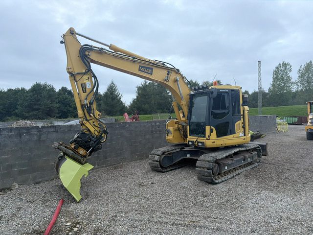Komatsu PC138US bæltegraver set skråt forfra med bommen strakt ud, tiltrotator og limegrøn skovl, foran en mur af betonblokke