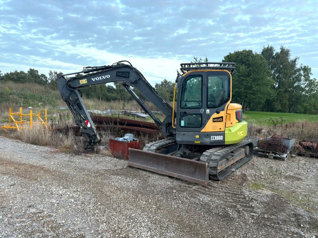 Volvo ECR88D minigraver på grusplads med dozerbladet forrest, kabinen og dørsiden mod kameraet, bommen svunget ud til venstre med stik og kobling, ECR88D-decal på hækken