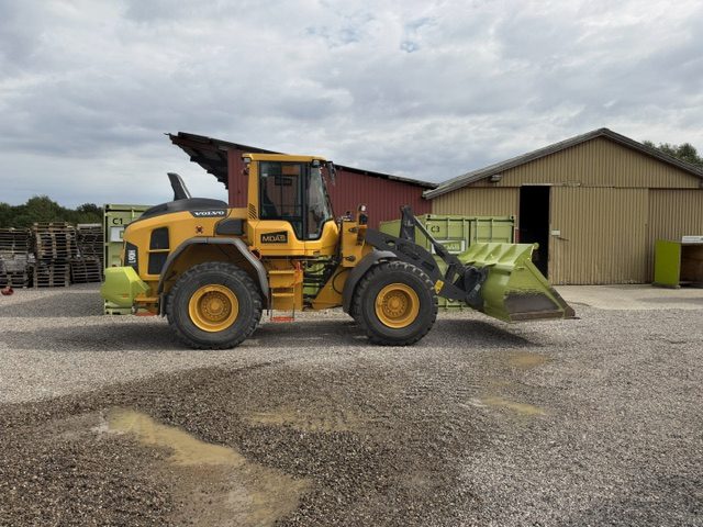 Volvo L 90 H gummihjulslæsser set fra siden med skovlen på jorden, foran grønne containere og en rødbrun bygning